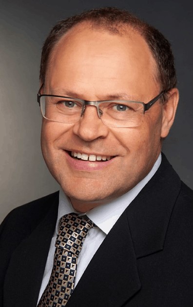 A picture of Eddy Rempel, a white man with brown hair and glasses, wearing a suit and tie. 