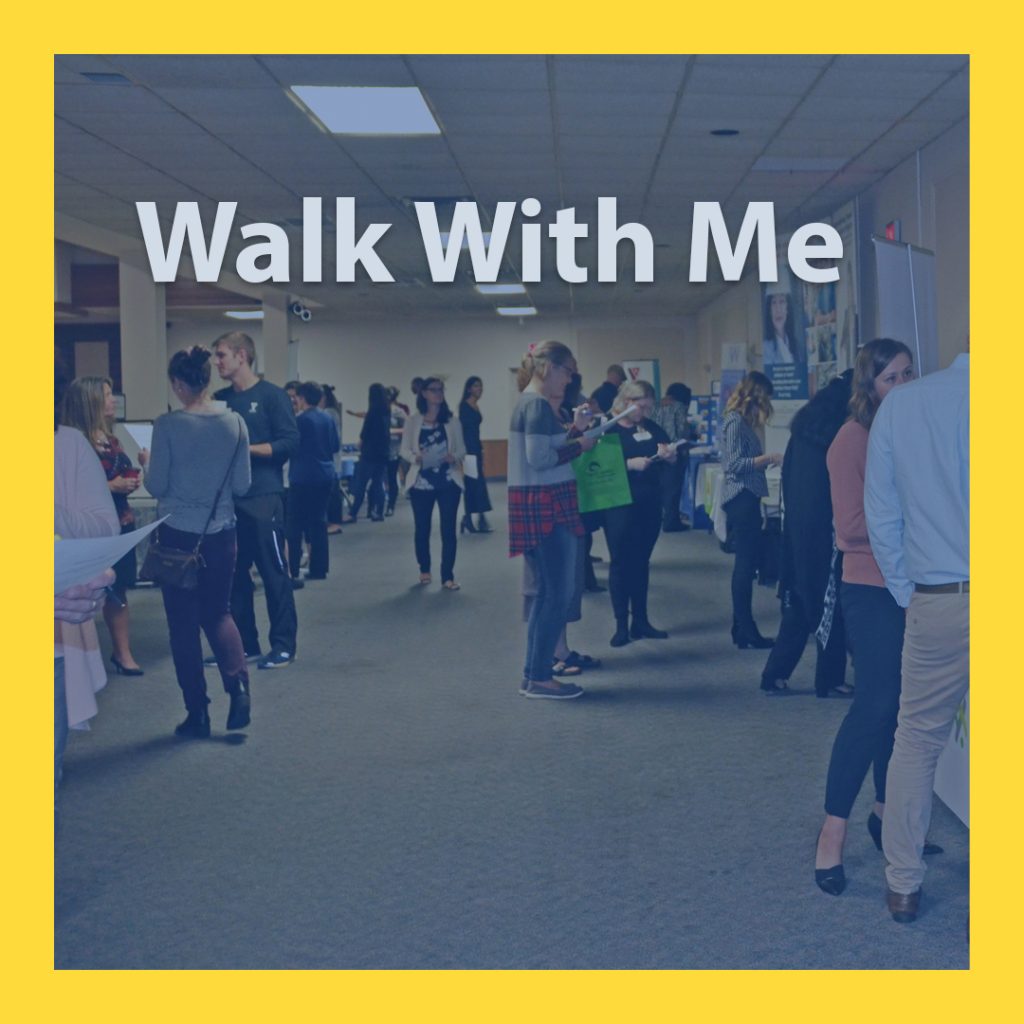 A group of professionals standing and talking at tables with banners set up next to them. There is text that reads Walk With Me. 