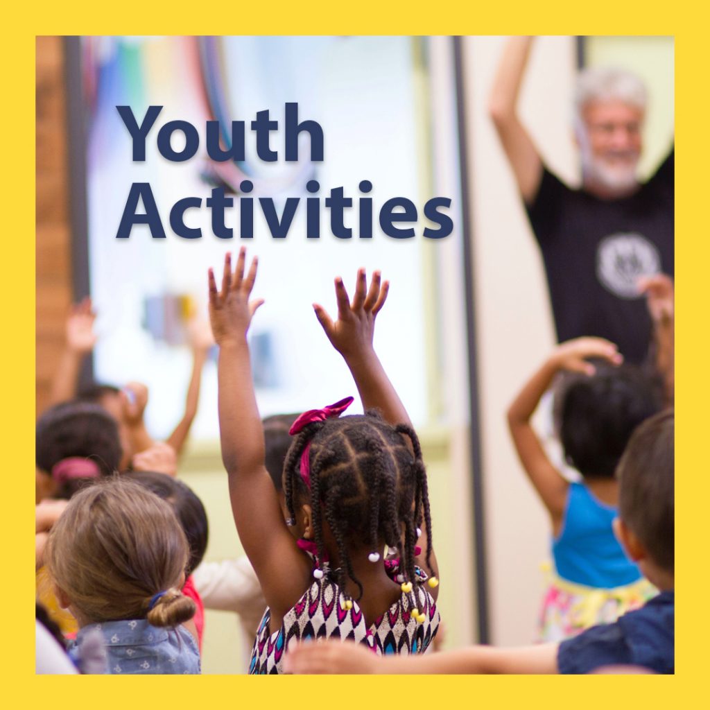 A group of young, multicultural children with their hands raised in the air, following the movement of an adult at the front of the room. There is text on the image that says Youth Activities. 
