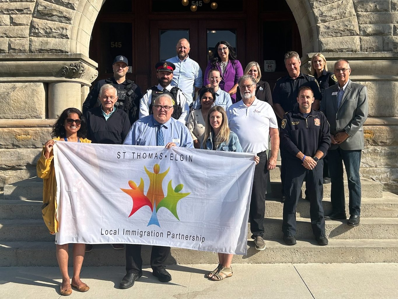 Community members and local officials holding St. Thomas-Elgin Local Immigration Partnership banner on steps of St. Thomas City Hall during flag raising and proclamation.