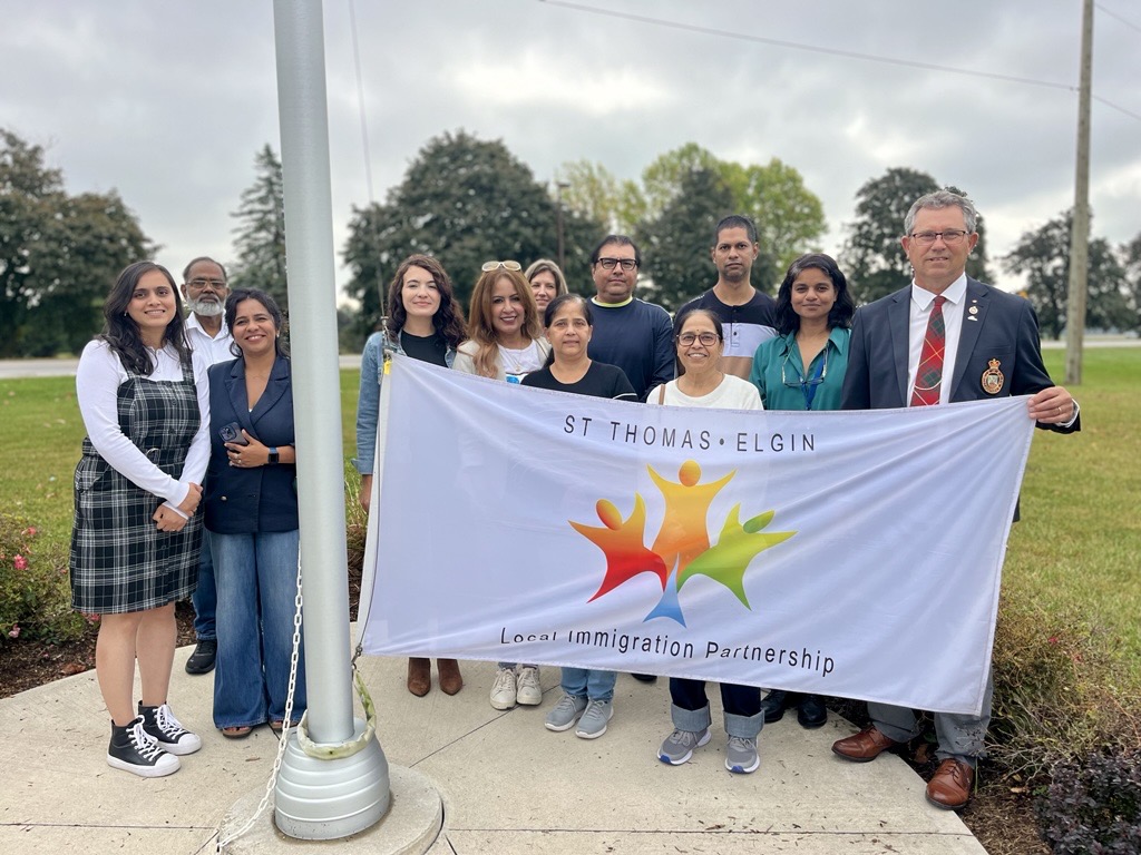 Group holding St. Thomas-Elgin Local Immigration Partnership banner during Elgin County Newcomer Open House, near flagpole.